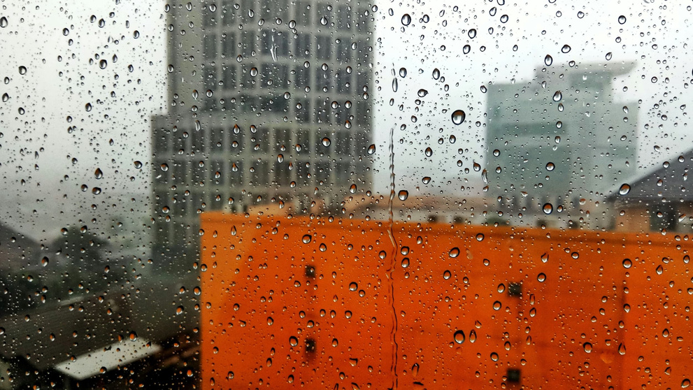 Raam met regendruppels, daarachter wazig stadsbeeld met grijze en oranje gebouwen onder grijze bewolkte lucht