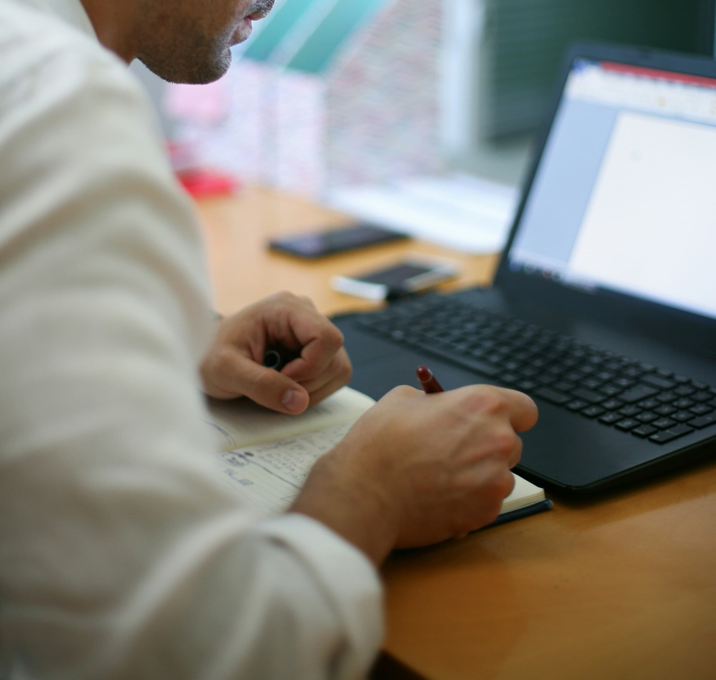 Handen schrijven in een blauw notitieblok met rode pen; op het bureau een open laptop en twee smartphones, over-de-schouder perspectief in kantooromgeving.